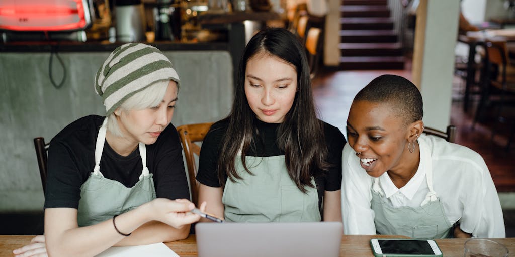 A cozy coffee shop scene featuring two friends sitting at a rustic wooden table, laptops open, and steaming mugs of coffee in hand. The warm lighting creates an inviting atmosphere, with a chalkboard menu in the background displaying various coffee options. The focus is on the interaction between the friends as they discuss ideas, with one of them typing on their laptop, symbolizing the concept of a 'Coffee Chat'.