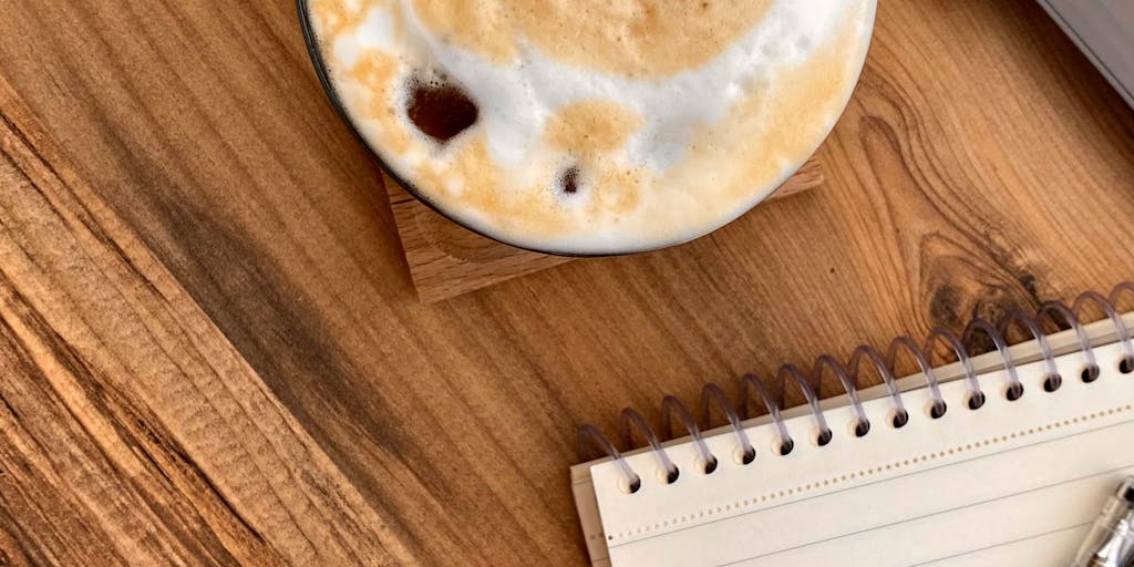 An overhead view of a desk with a laptop open to the Klenty pricing page, a cup of coffee, and a notepad filled with brainstorming ideas. The scene is well-lit, with a plant in the background to add a touch of freshness.