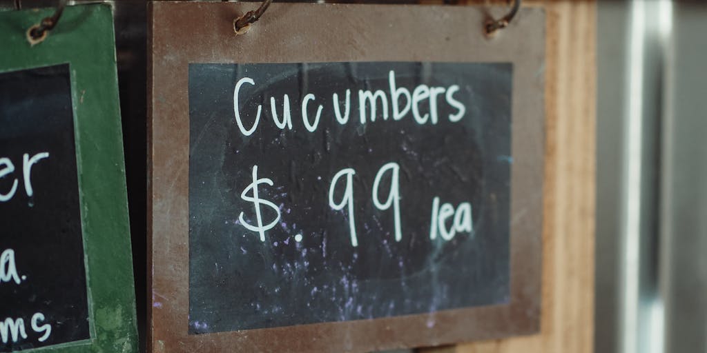 A close-up shot of a chalkboard with handwritten pricing tiers for a fictional food delivery service called 'Yamm'. The board is surrounded by colorful ingredients like fresh vegetables, fruits, and spices, creating a vibrant and appetizing atmosphere.