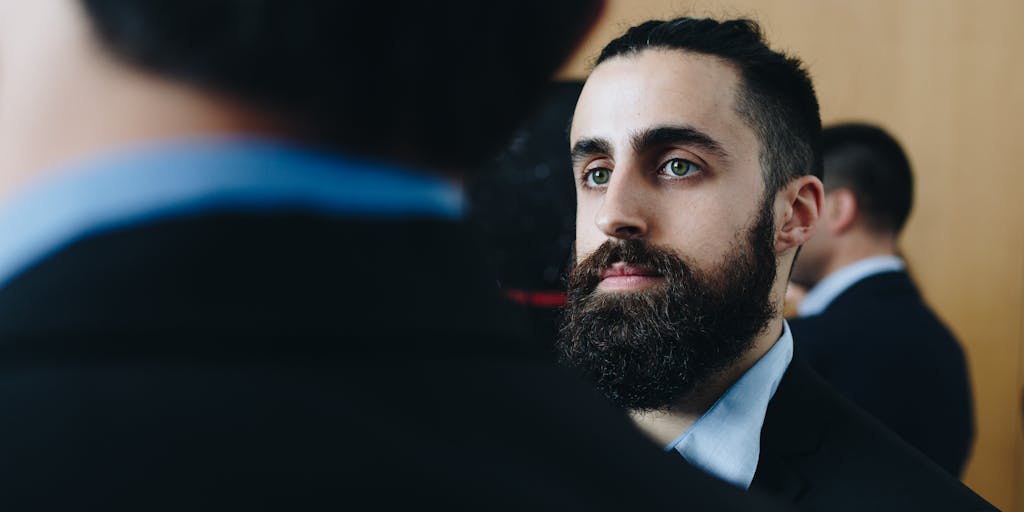 A dynamic shot of a person confidently networking at a business event, holding a smartphone displaying their LinkedIn profile. The background features other professionals engaging in conversation, emphasizing the importance of having an optimized profile for making valuable connections in sales.