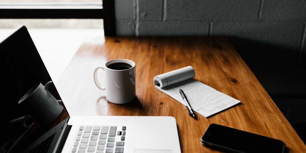 An overhead view of a workspace featuring a smartphone displaying the LinkedIn app, a cup of coffee, and a notepad filled with email templates. The scene is well-lit, with a focus on the smartphone, suggesting a casual yet productive environment for gathering emails.